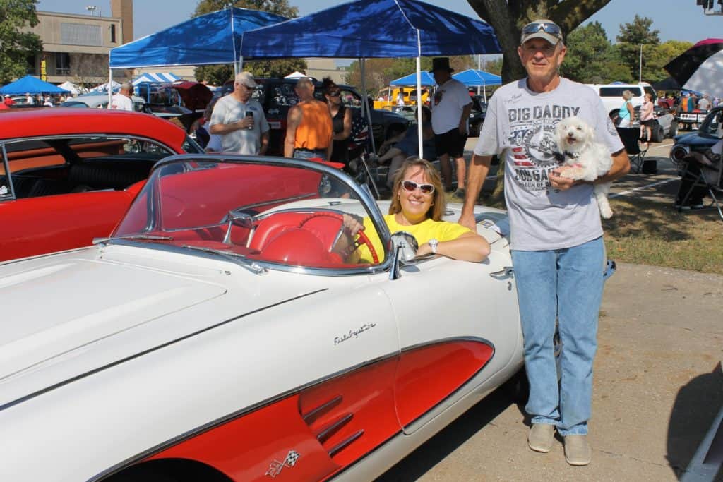 Bettie Truitt sitting in white & orange Corvette, Ron Williams standing next to car holding white dog
