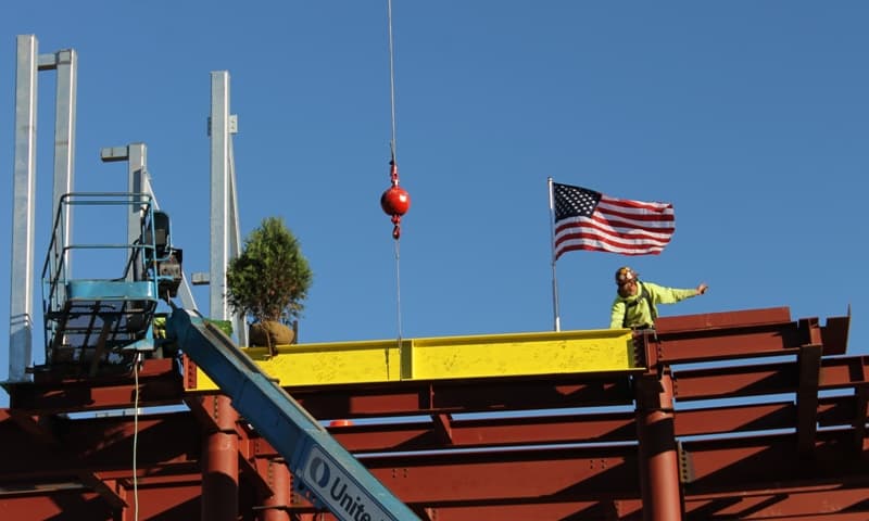Closeup of yellow beam with evergreen & American flag and worker on top of red steel framework of new 2-story building