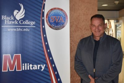 Black Hawk College Veteran's Coordinator Thomas Reagan standing next to a red, white, and blue banner for the Military Students and Veterans Club