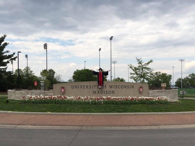 person in graduation gown sitting on University of Wisconsin Madison sign, surrounded with red and white flowers