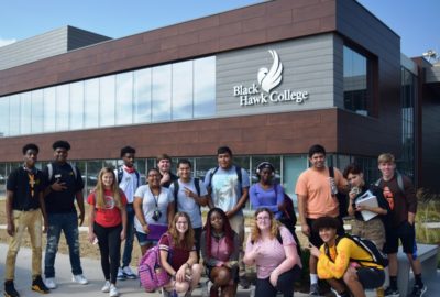 Large group of students pose in front of campus building on a sunny day