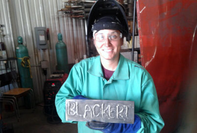 smiling woman in welding gear holding a welded plaque that says her last name, BLACKERT