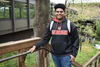 smiling male college student standing at the top of a staircase on campus with trees in the background