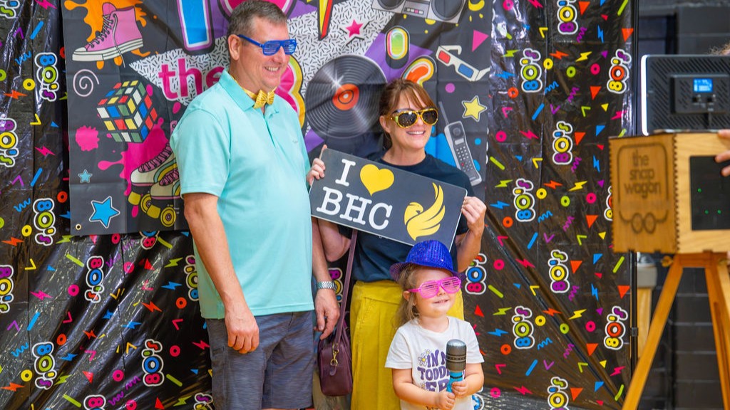 2 adults and 1 child standing in front of colorful 80-themed backdrop holding i heart bhc sign.