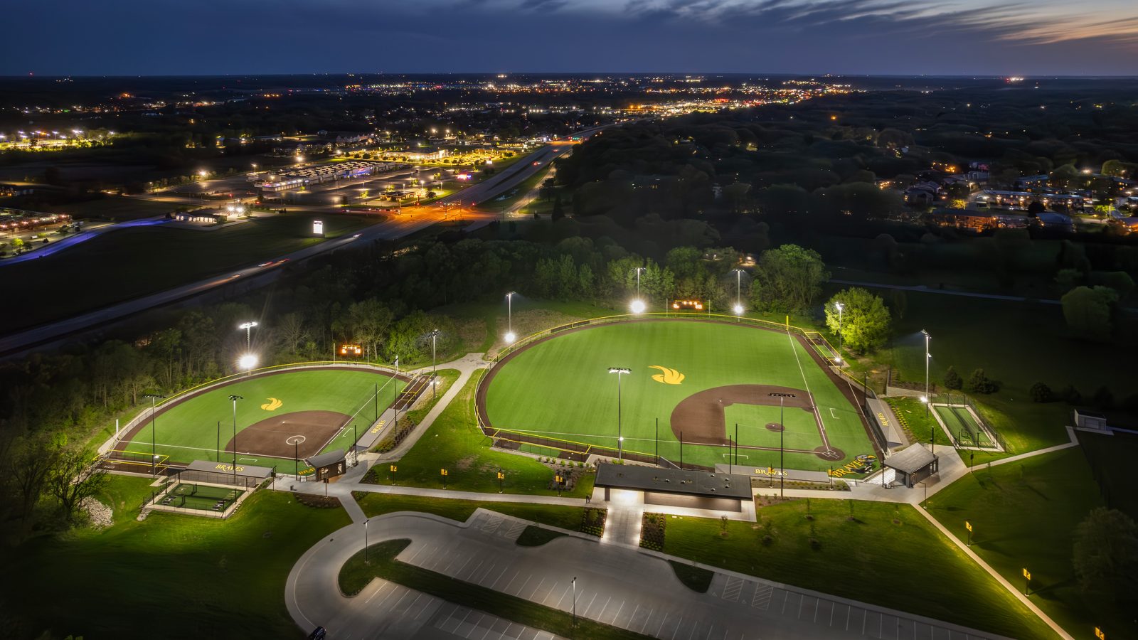Athletic complex at night featuring two baseball/softball diamonds and a glowing city behind it.