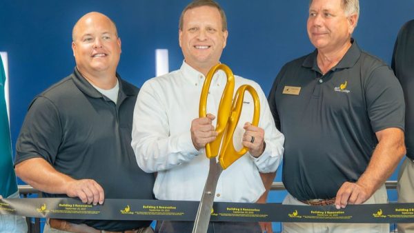 3 people in a line holding a ribbon with one man cutting ribbon with oversized scissors.