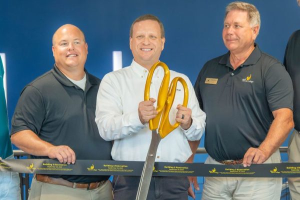 3 people in a line holding a ribbon with one man cutting ribbon with oversized scissors.
