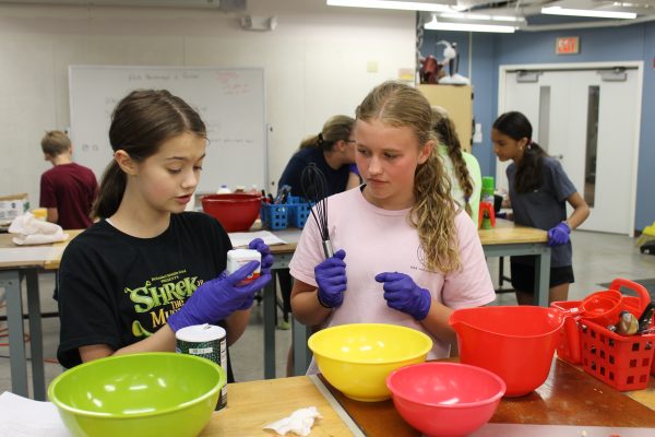 Over 500 students explore passions at college for kids Two girls in the kitchen. One, in a pink shirt, is holding a whisk, while the other in a black shirt is holding salt.
