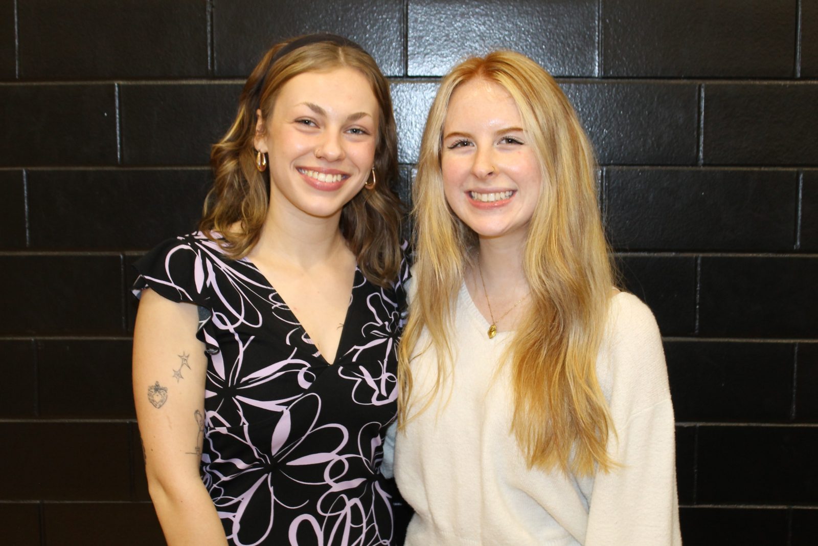 2 women standing in front of black wall and smiling at camera.