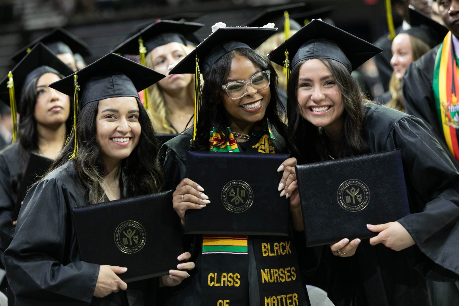 A group of graduating women smile, holding their diplomas at a commencement ceremony.