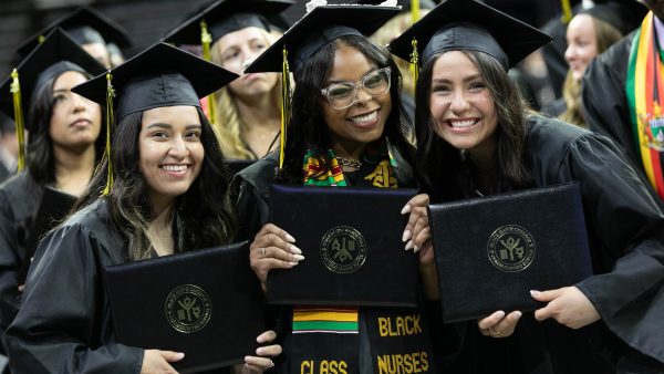 A group of graduating women smile, holding their diplomas at a commencement ceremony.
