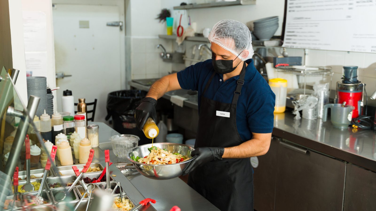 Food protection manager Latin cook wearing black gloves, apron, hairnet, and face mask pouring dressing on a healthy salad inside a restaurant kitchen