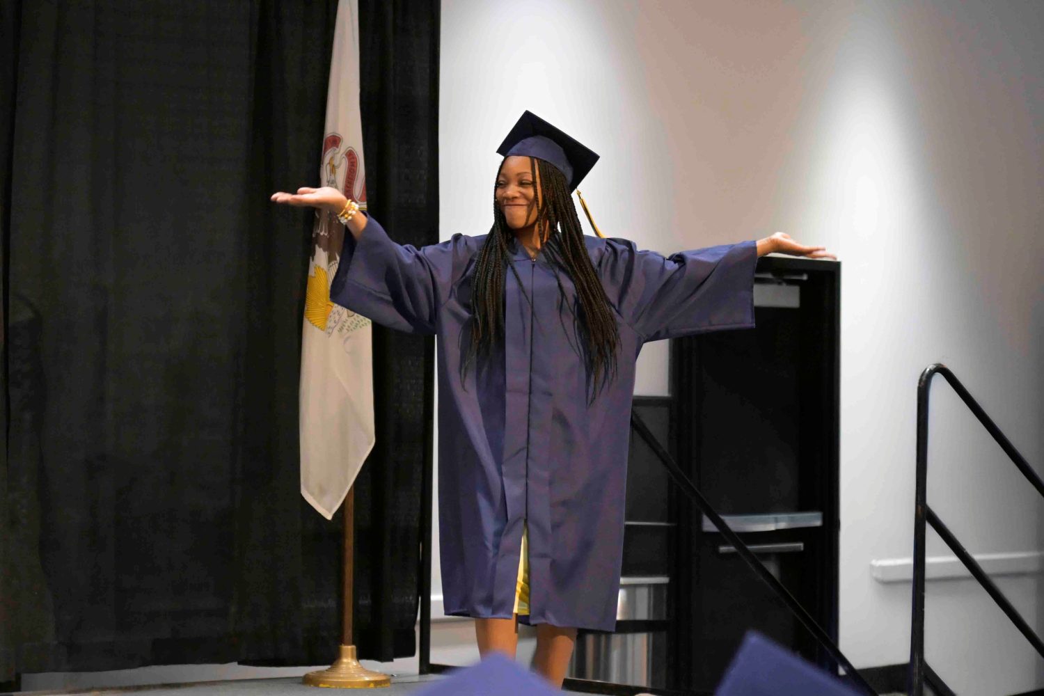 Grad in cap and gown with arms raised standing on a stage.
