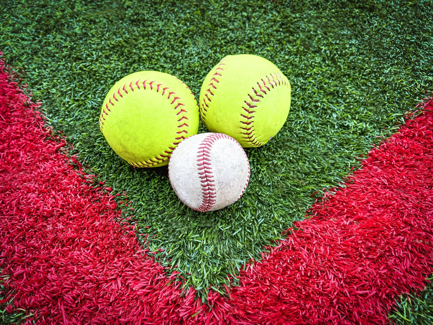 Two neon yellow softballs and one baseball lay on a green turf field. Red outline indicates batting area. Overhead views.