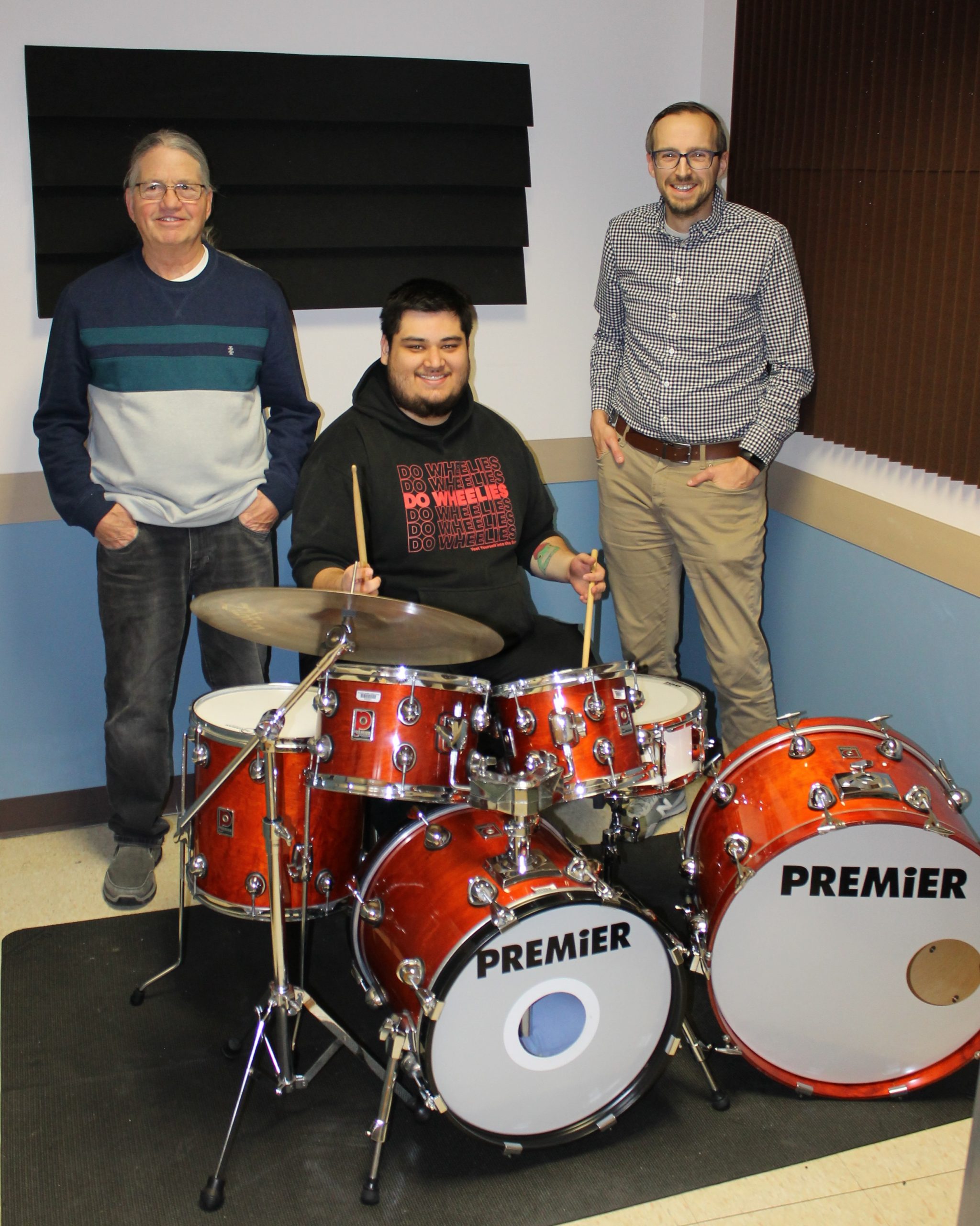 One person sitting at drum kit with 2 people standing.