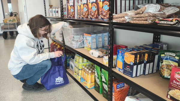 First generation and underrepresented students A woman wearing a gray hoodie and blue jeans is crouching down while selecting items from a shelf stocked with food and drinks. They are placing the items into a blue reusable bag with the logo