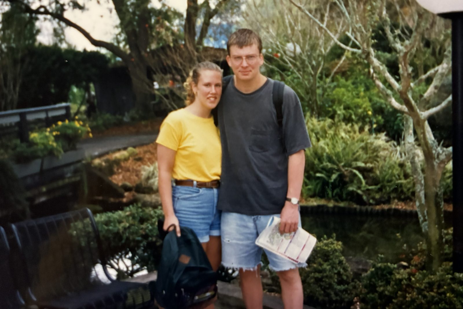 Woman and man standing outside in front of trees, bench and landscaping.