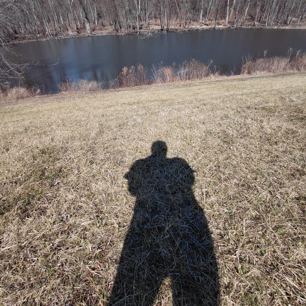 Shadow of person standing on dead grassy area next to a river or creek.