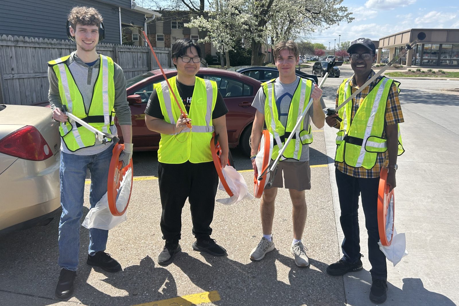 4 people wearing safety gear standing in parking lot with trash bags and grabbers to pick up trash.