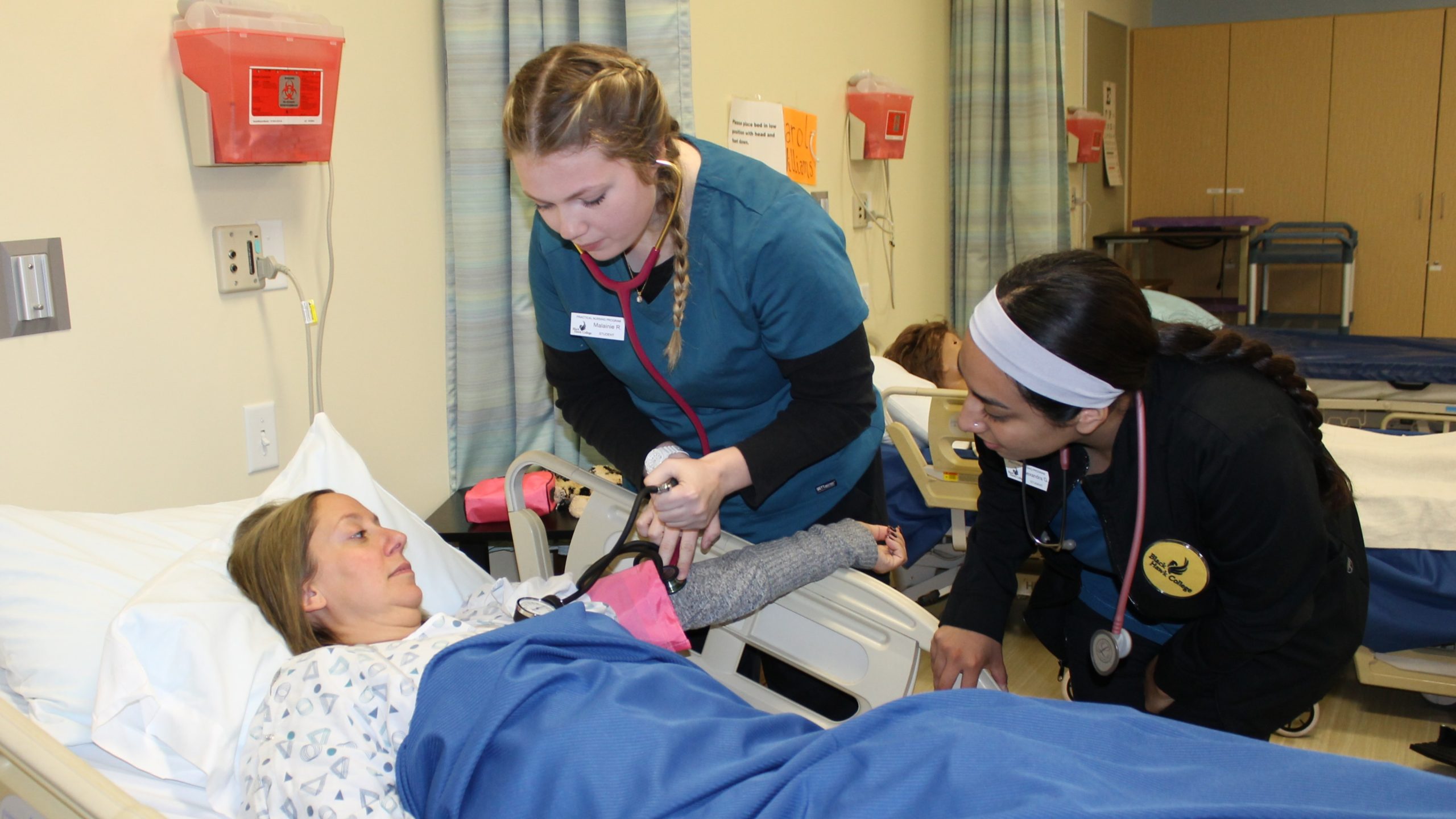 2 practical nursing students taking blood pressure of patient in a hospital bed.