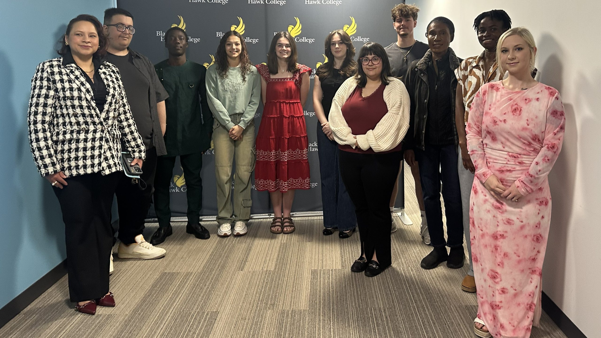 Qc campus students join phi theta kappa honor society - spring 2026 11 people smiling at the camera and standing in a hallway with black hawk college backdrop.