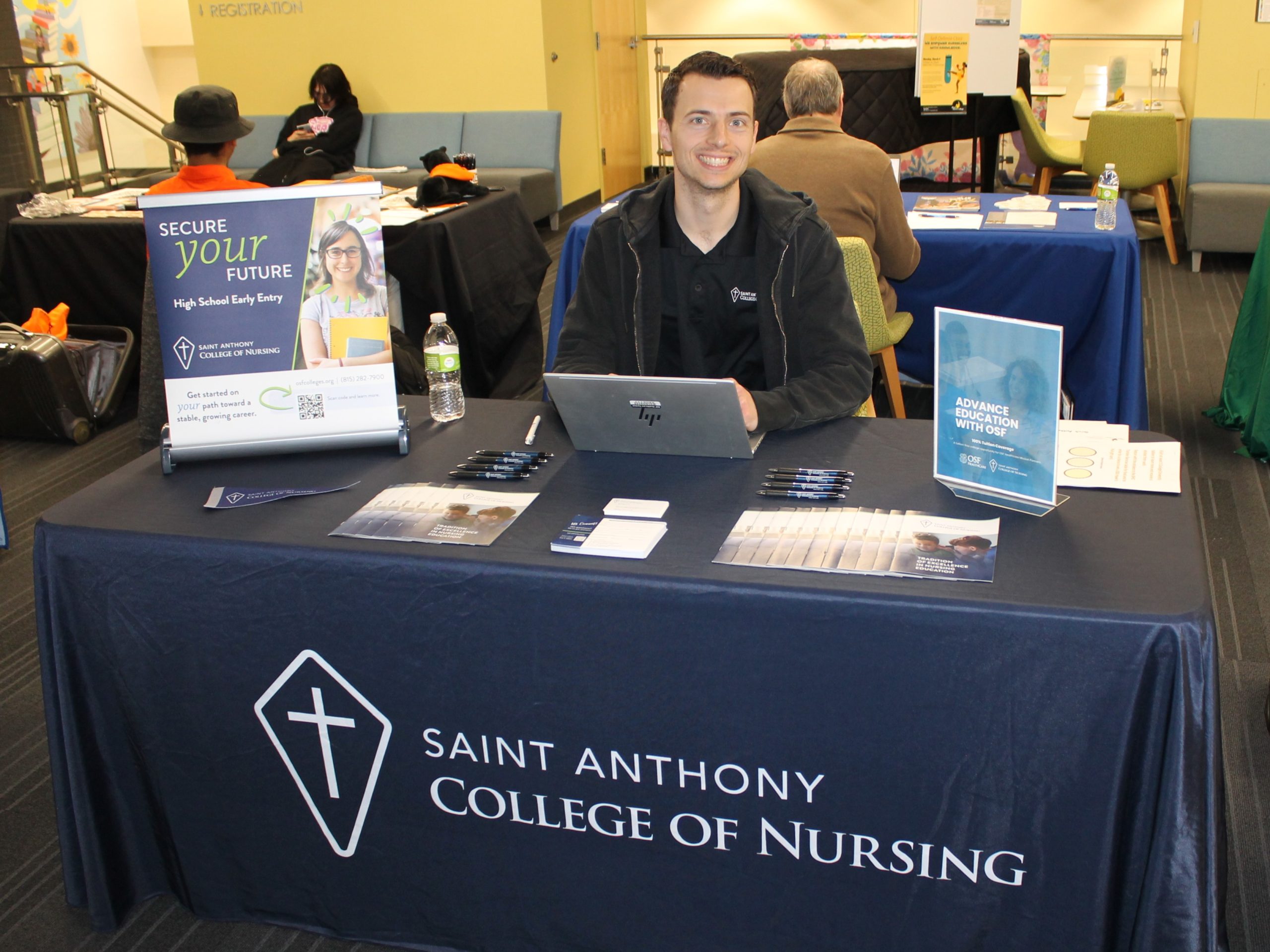 Saint anthony college of nursing visit Person sitting at table covered by tablecloth and college promotional materials.
