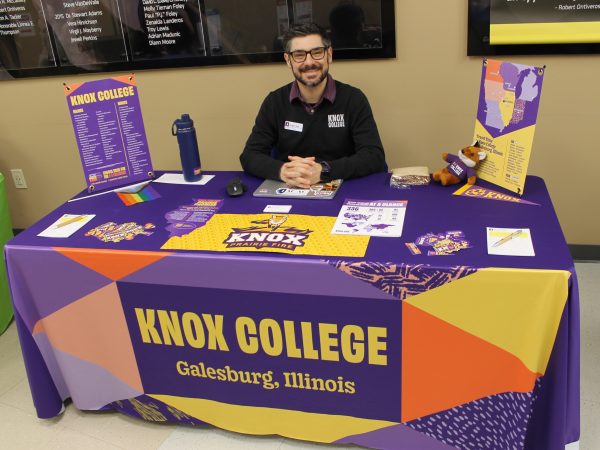 Person sitting at table covered by tablecloth and college promotional materials.