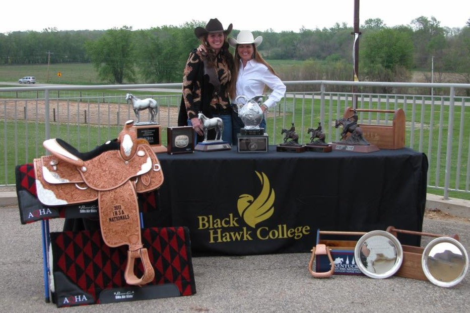 2 women standing outdoors behind a table covered with awards and saddle in front of table.