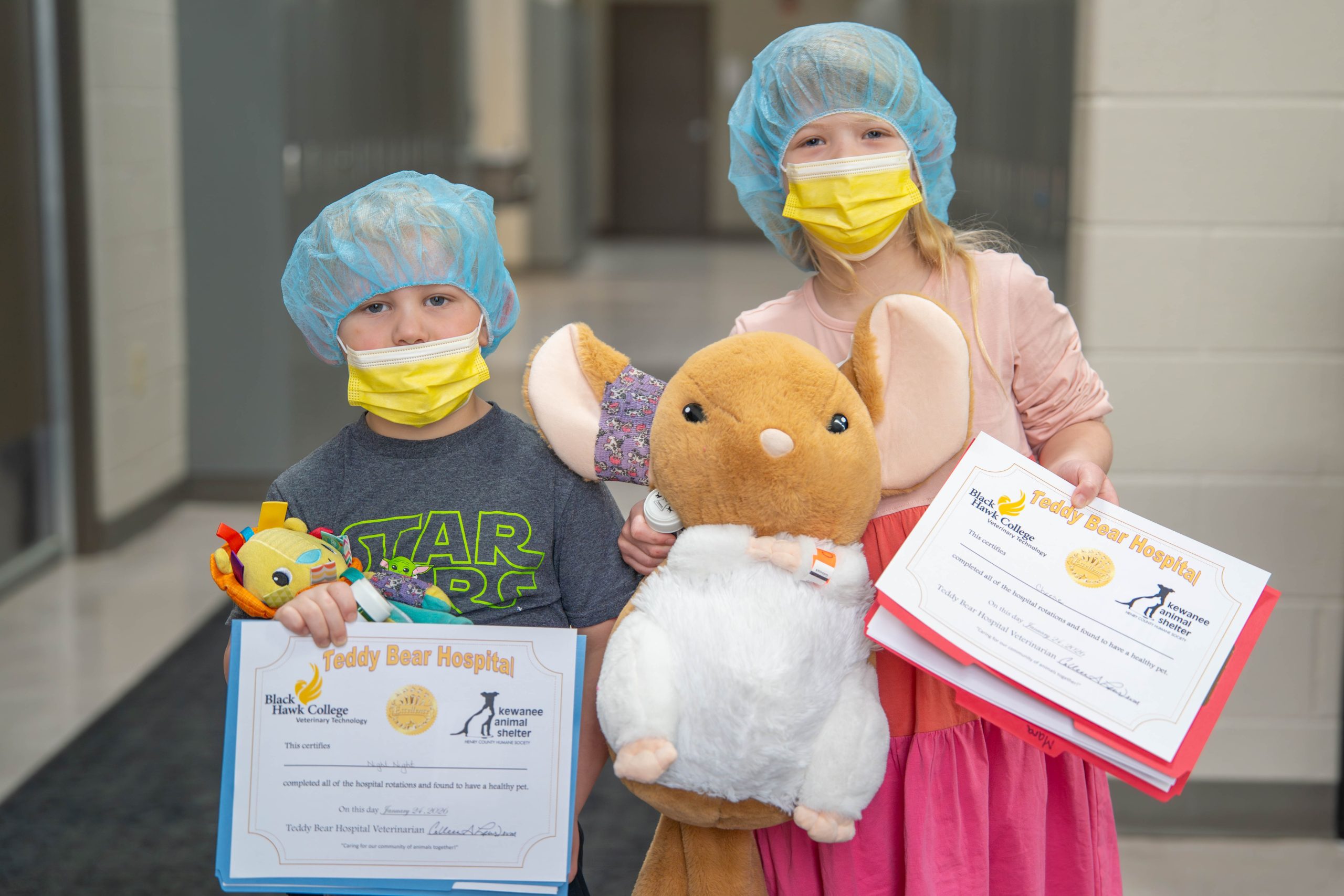 2 children wearing medical face masks and hair nets looking at camera and holding stuffed animals and certificates.