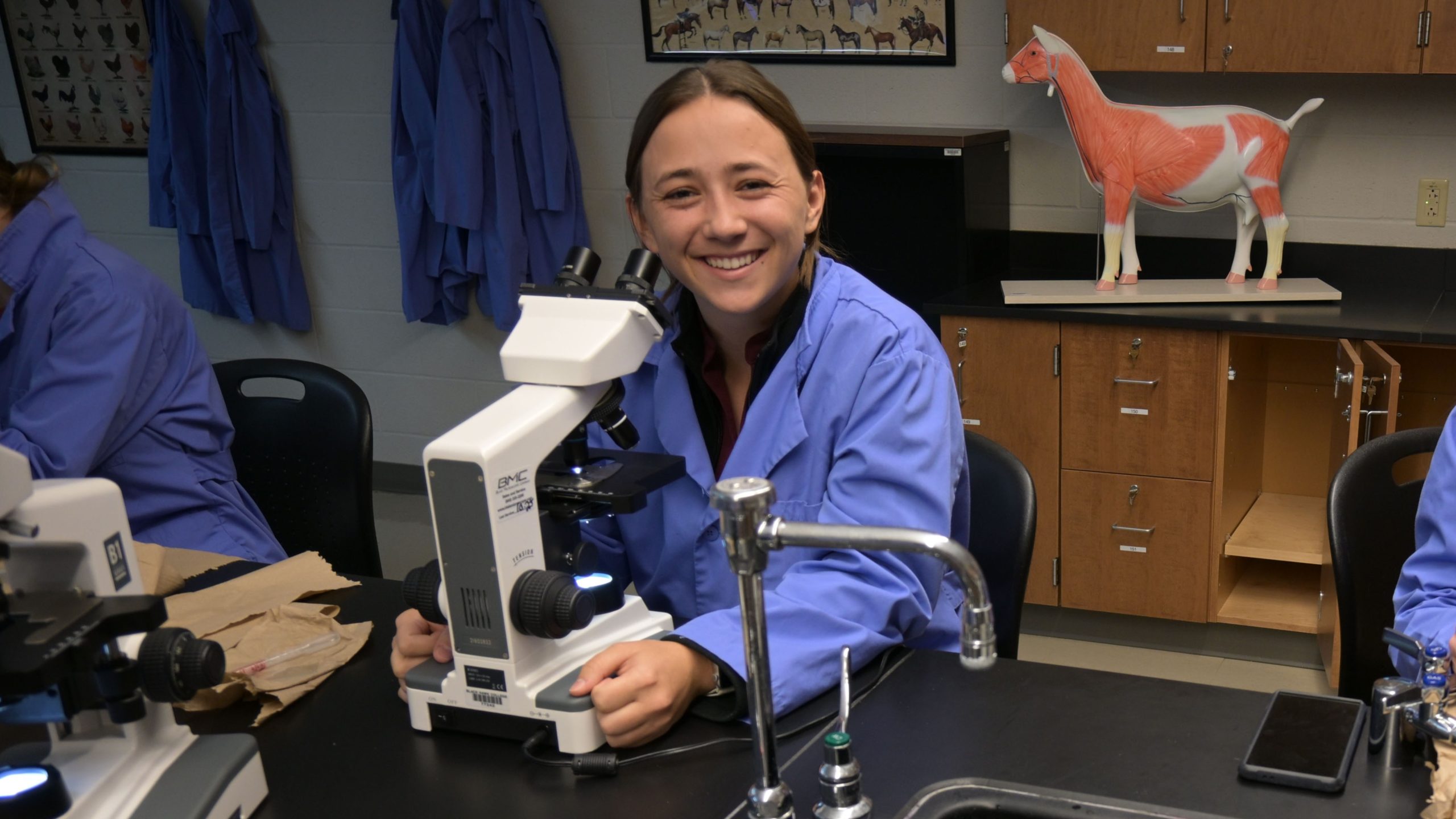 Veterinary technology open house Student in lab coat sitting at classroom lab table with microscope.