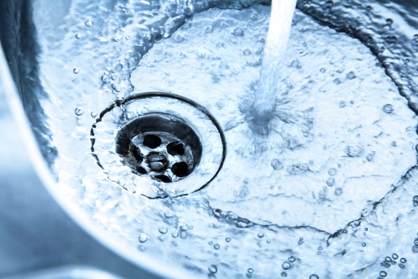 Close up of a stainless steel kitchen sink with running water. The faucet is turned right up. A strong water jet flows into the basin. Small bubbles and swirls are seen. The water flows through the open drain. Sharp and detailed image. Blue toned