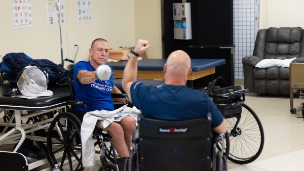 Two individuals in wheelchairs engage in a throwing activity within a clinical or rehabilitation setting.