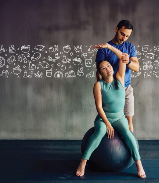 Woman sitting on exercise ball stretching with man standing behind her assisting.